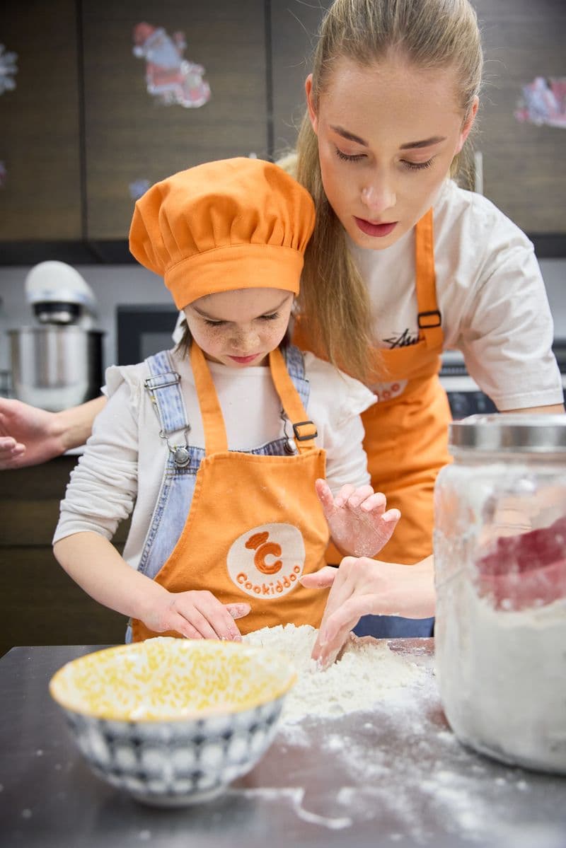 A child in a chef hat kneads dough with a coach guiding her hands